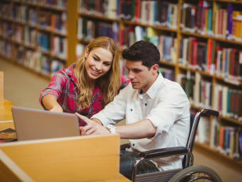 Biblioteka, mężczyzna na wózku inwalidzkim przy komputerze obok pochyla się kobieta